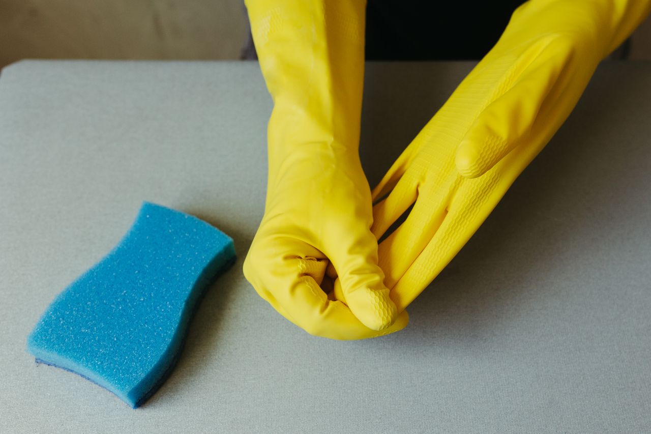 services-01 Close-up view of hands in yellow gloves beside a blue sponge on a gray surface.